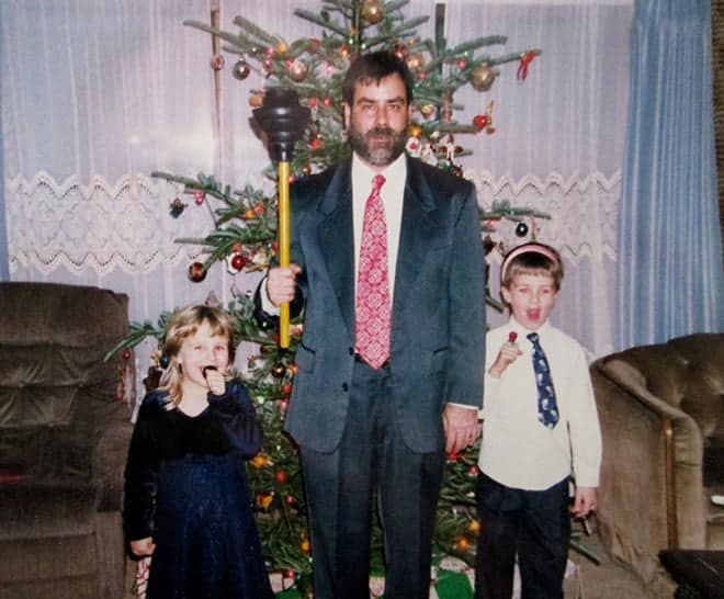 An awkward family christmas photo of a dad holding a plunger while kids stand by a tree
