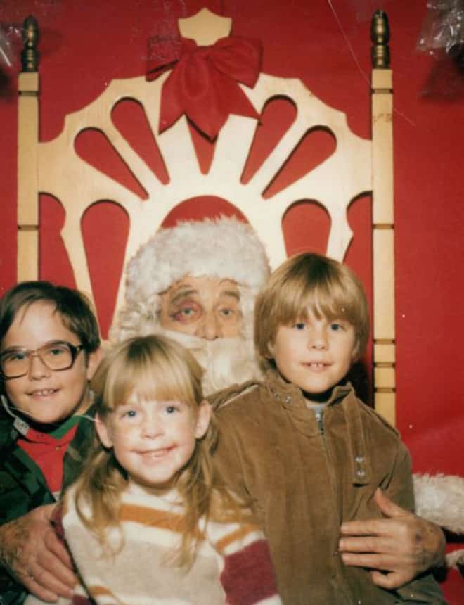 An awkward family christmas photo of two kids sitting on a Santa who has a black eye.