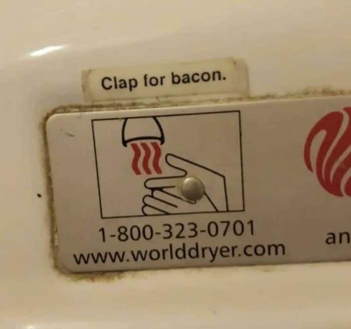 A creative vandalism sticker on a hand dryer changing the instructions to "Clap for bacon".