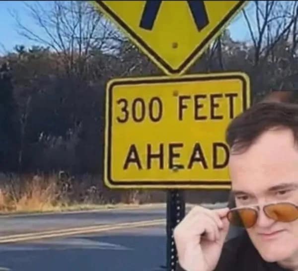 Quentin Tarantino looking interested next to a road sign reading three hundred feet ahead.