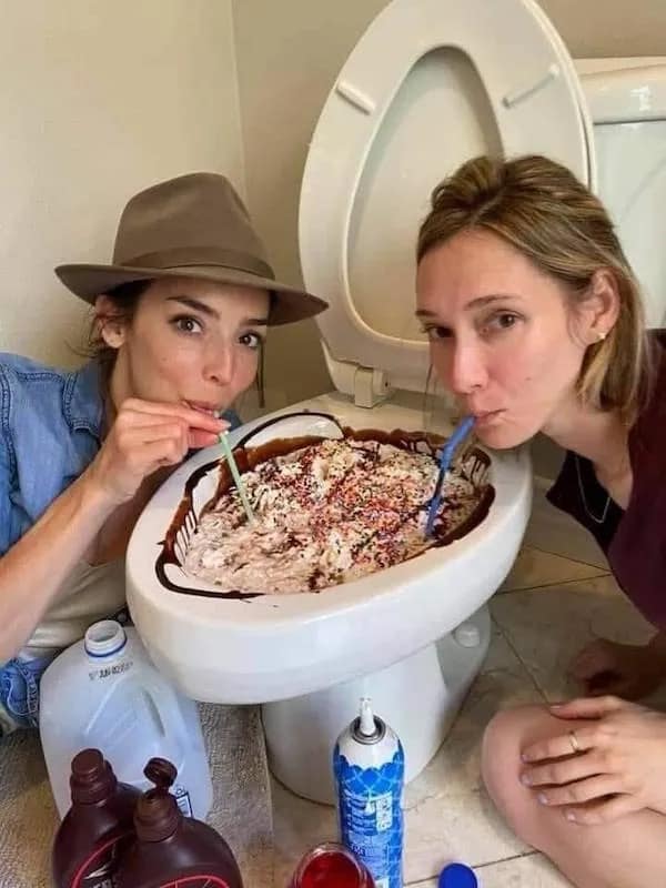 Two women drinking milkshake out of toilet bowl with straws.
