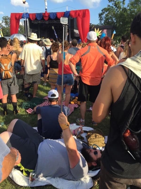 Man lying on grass at concert using selfie stick to view stage.