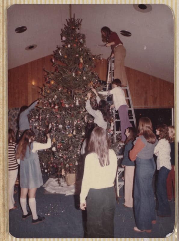 A retro christmas photo of a family decorating a tall tree on a ladder.