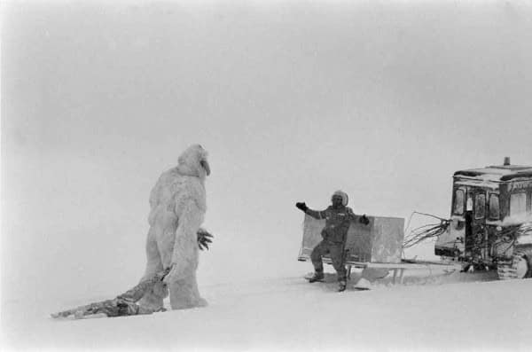 A Star Wars BTS photo of a Wampa dragging a Luke dummy while a crew member directs in the snow.
