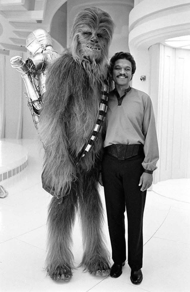 A Star Wars behind the scenes photo of Billy Dee Williams smiling next to Chewbacca carrying C-3PO