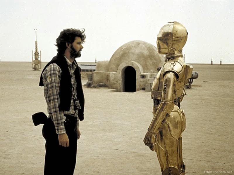 A Star Wars BTS photo of George Lucas talking to C-3PO in the desert of Tatooine.