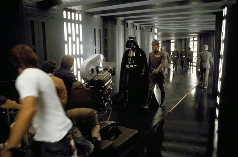 A Star Wars behind the scenes photo showing the camera crew filming Darth Vader walking down a hallway.