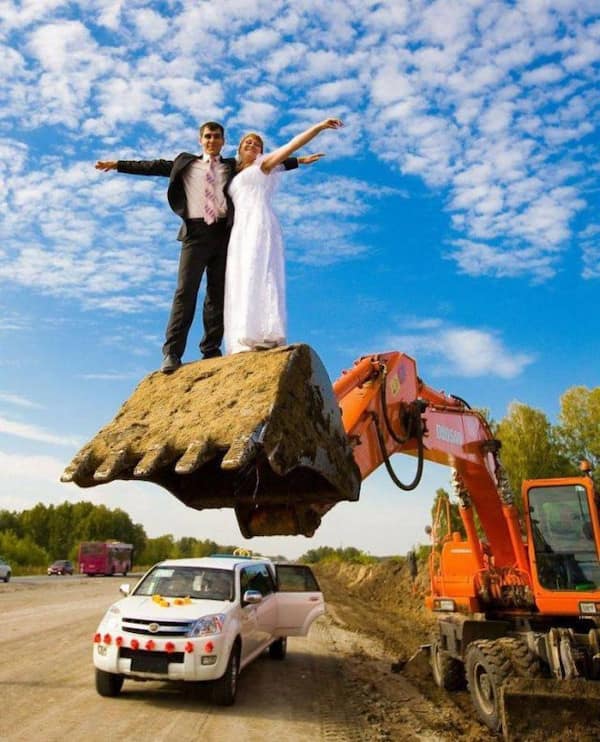 Bride and groom posing like Titanic on dirty excavator bucket above car.