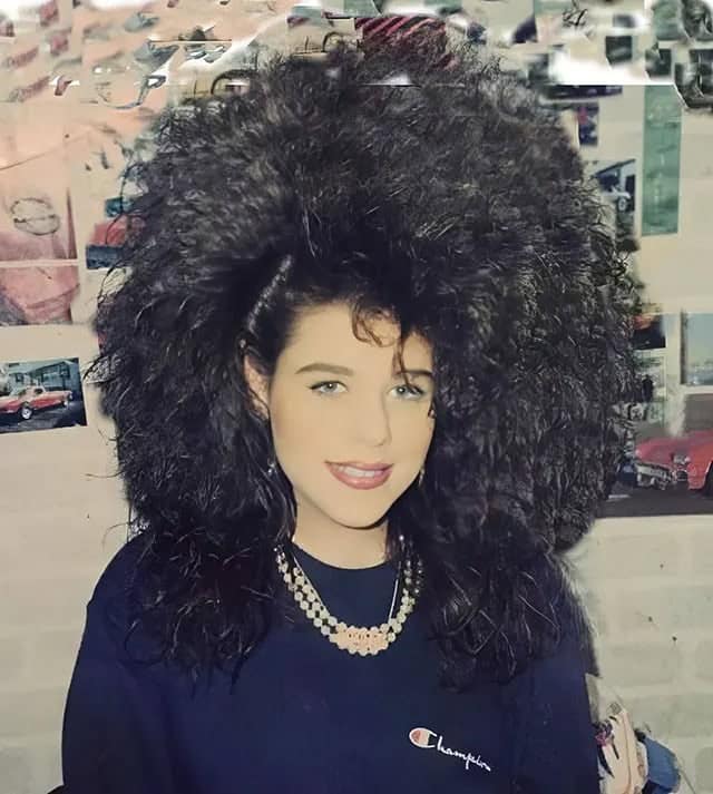 A woman with a massive, dense black 80s haircut that frames her face like a helmet.