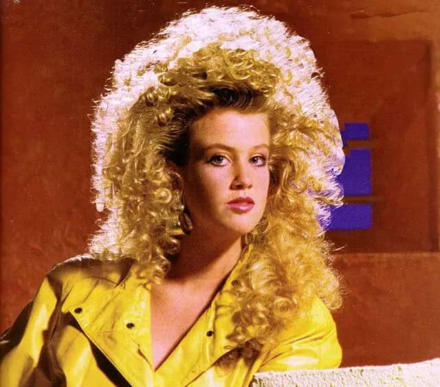 A blonde woman wearing a yellow jacket with a curly, backlit 80s haircut.