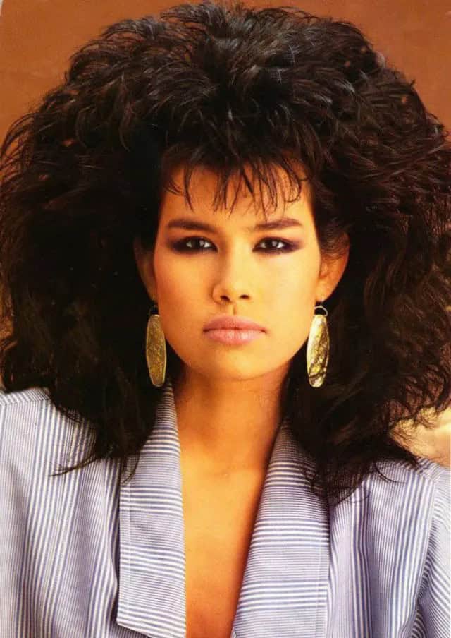 A woman with a wide, dark 80s haircut featuring heavy bangs and feathered sides.