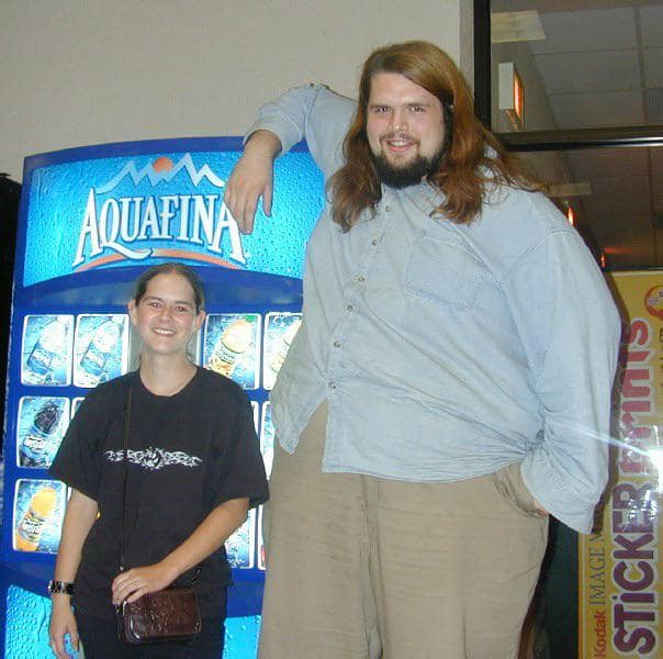 An absolute unit of a tall man leaning on a vending machine, towering over the woman standing next to him.