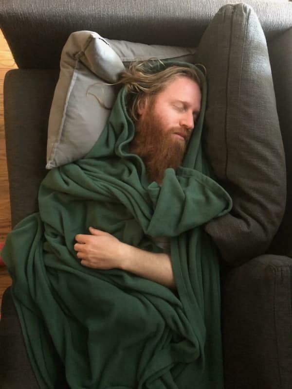 Bearded man sleeping on a couch wrapped in heavy green fabric folds.