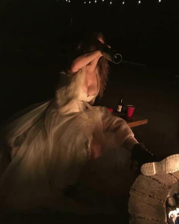 Woman in a gown drinking wine by a fire pit at night.