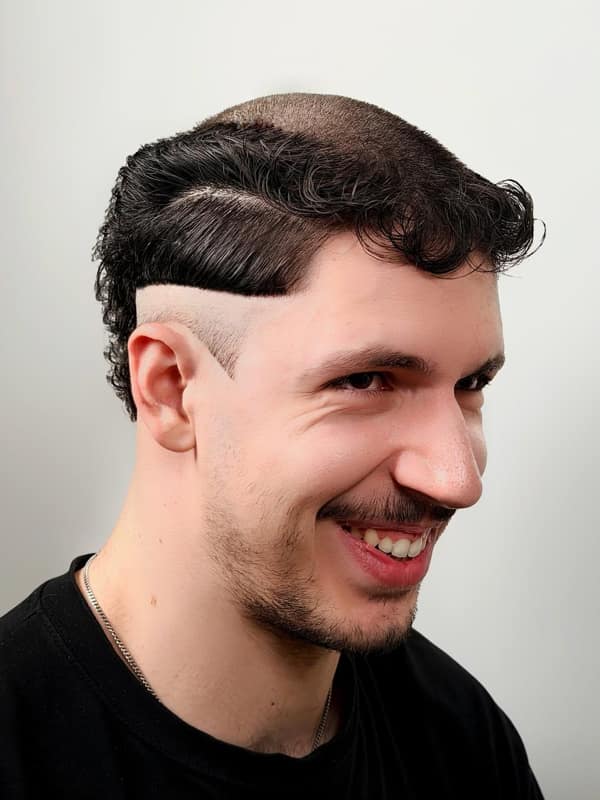 A man smiling with a haircut fail that is shaved on top with curly bangs and a mullet back.