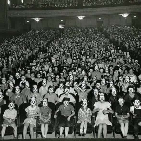 Large theater audience of children wearing identical smiling Mickey Mouse masks in black and white.