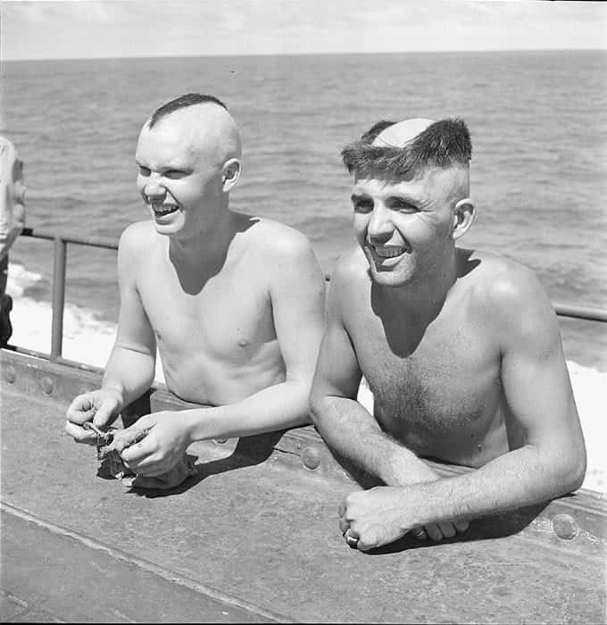 Two shirtless men smiling with hair shaved down the middle leaving tufts on the sides.