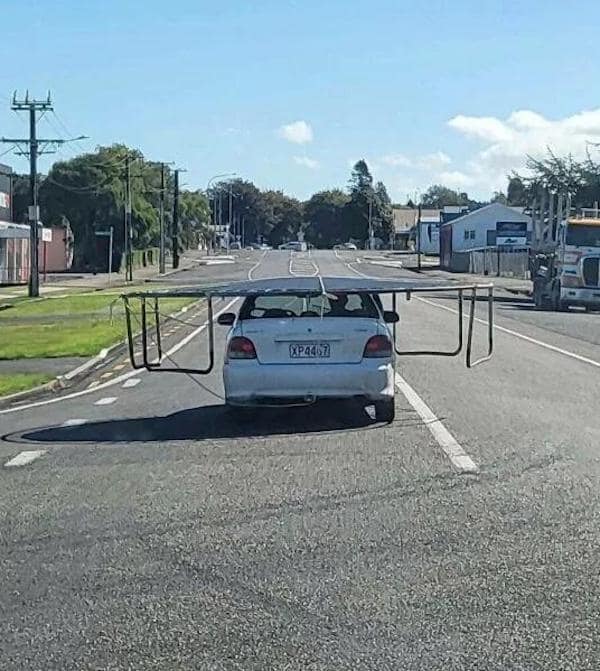 A driving fail showing a small white car transporting a giant trampoline frame on its roof.