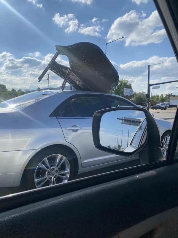 A bad driver photo of a sedan with a wicker bench seat strapped to the roof at a steep angle.