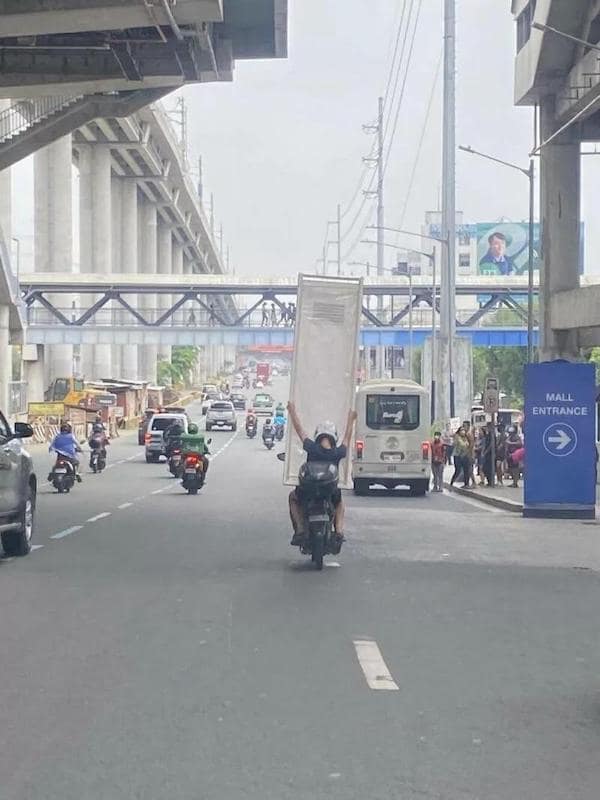 A bad driver photo of a motorcyclist carrying a full door vertically on their back in traffic.