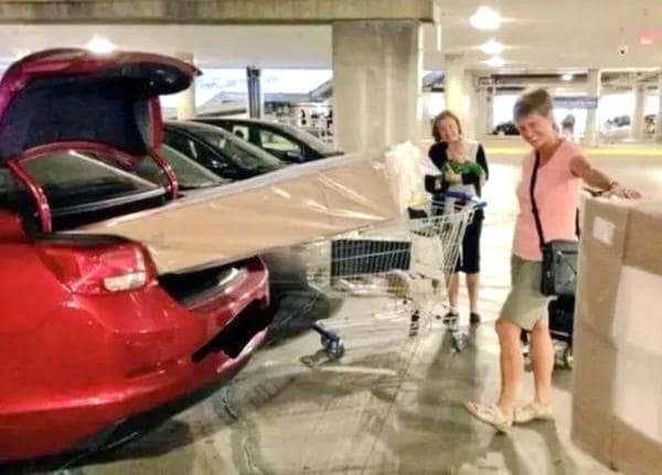 A driving fail showing a long box sticking out of a car trunk supported by a shopping cart.
