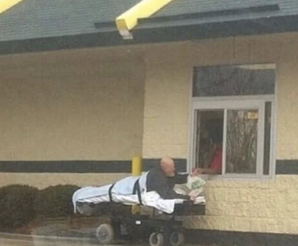 Patient on a hospital gurney ordering food at a drive-thru window.