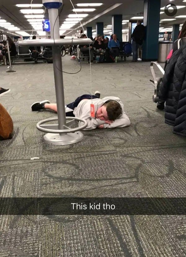Young boy sleeping on an airport carpet with a phone charger cable dangling over his face.