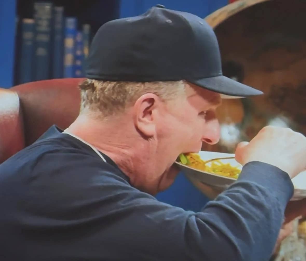 Man in a navy baseball cap aggressively shoveling food into his mouth from a bowl.