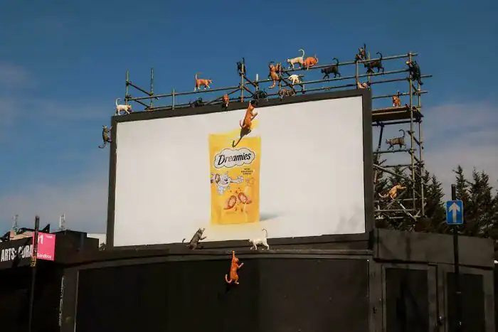 A funny print ad billboard for Dreamies cat treats with realistic cat mannequins climbing the structure to reach the pack.