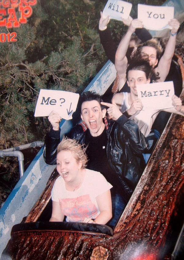 A funny rollercoaster picture of a marriage proposal happening on a log flume with friends holding signs.