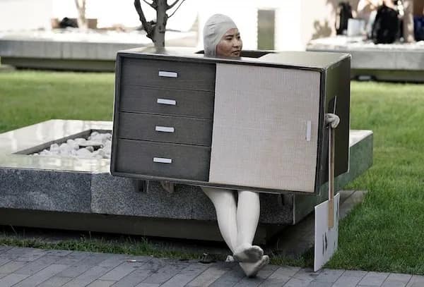 A hilarious meme photo of a person wearing a realistic wooden dresser costume sitting on a bench.