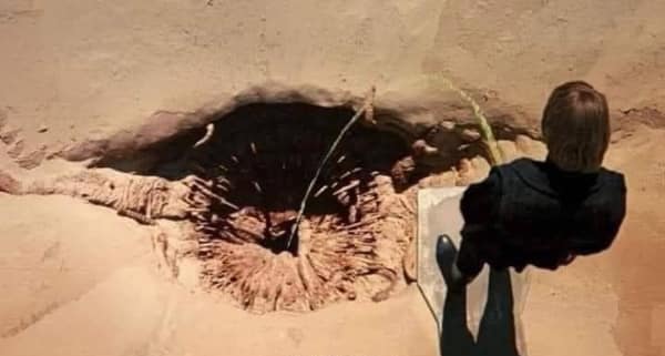 A movie meme top-down view of a character looking into a giant, toothy desert pit resembling the Sarlacc from Star Wars or the creatures in Dune.