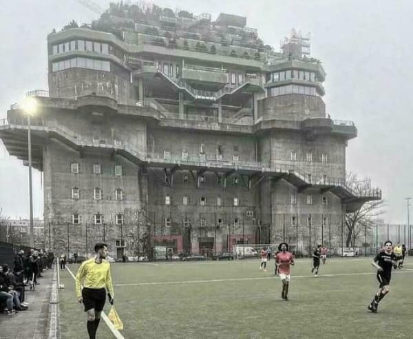 A massive concrete sinister building overlooking a soccer field resembling a fortress or bunker.