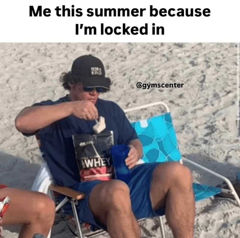 Man on a beach mixing protein powder in a shaker while sitting in a folding chair.