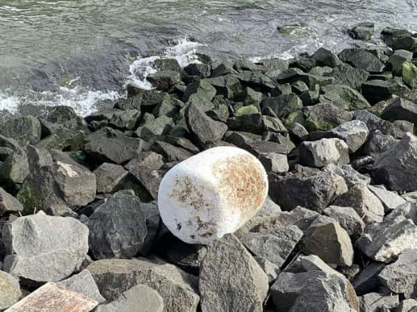 Large white cylindrical object washed up on rocky shore resembling a giant marshmallow.