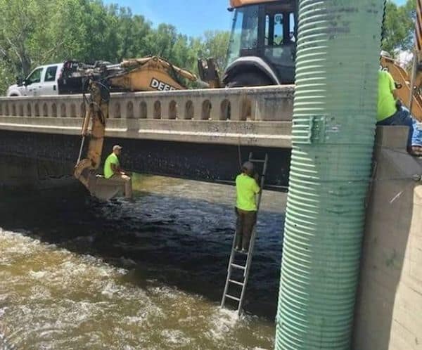 onstruction workers sitting in excavator bucket and on hanging ladder over river.