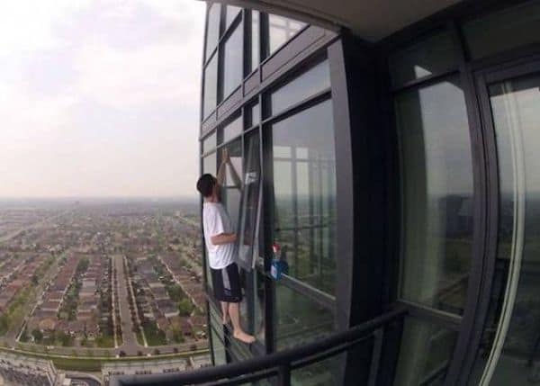 Man standing barefoot on high-rise building ledge cleaning window without safety gear.