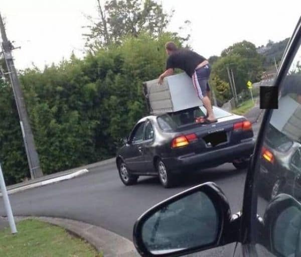Man standing on moving car trunk holding unsecured refrigerator on roof.