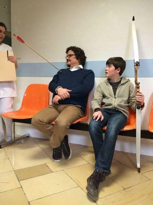 Man sitting in waiting room with arrow in chest next to boy holding bow.