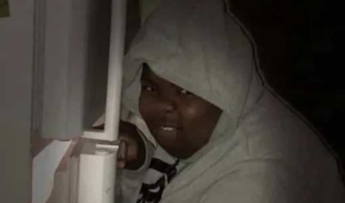 A funny low-light photo of a person in a grey hoodie peeking mischievously out from behind a refrigerator.