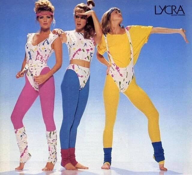 Three women modeling colorful Lycra spandex 80s fitness outfits with paint-splatter patterns and leg warmers.