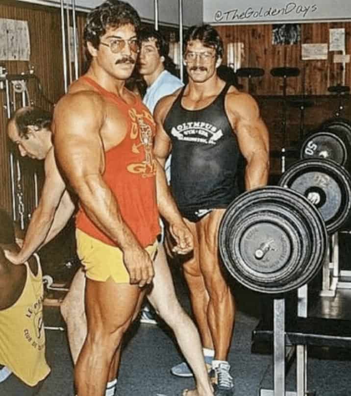 Two muscular men with mustaches and glasses training with barbells in a vintage gym setting.
