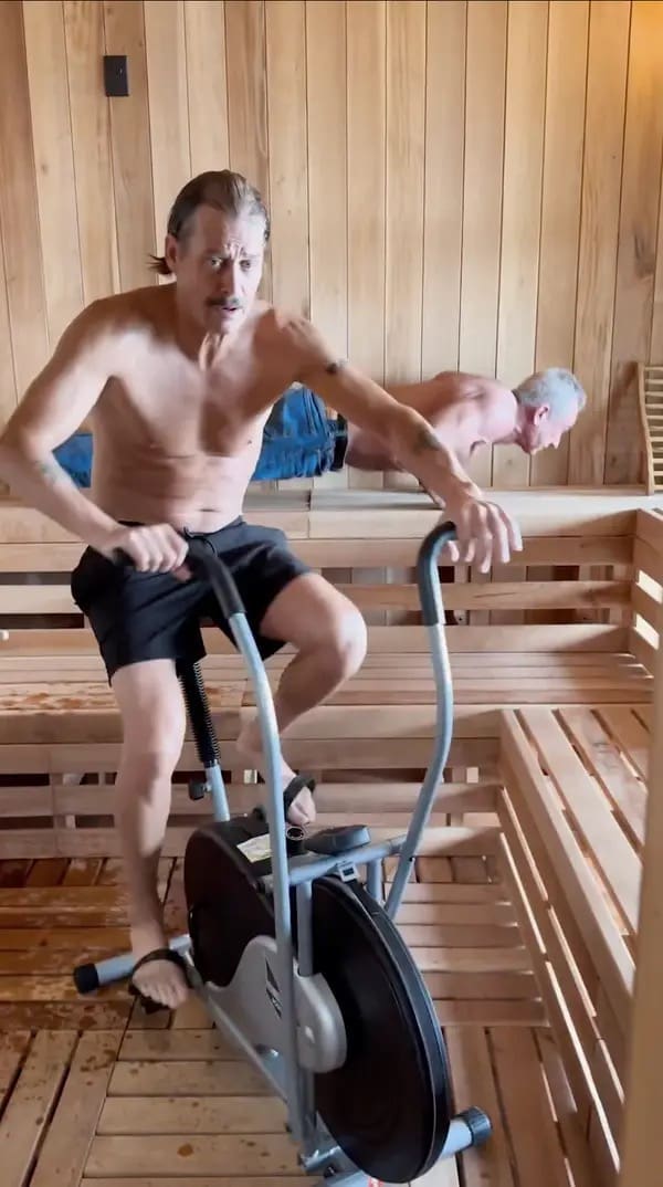 A chaotic RFK JR and Kid Rock workout photo showing Kid Rock pedaling the stationary bike in sandals while RFK Jr., still fully clad in denim jeans, aggressively does pushups on the wooden sauna benches in the background.