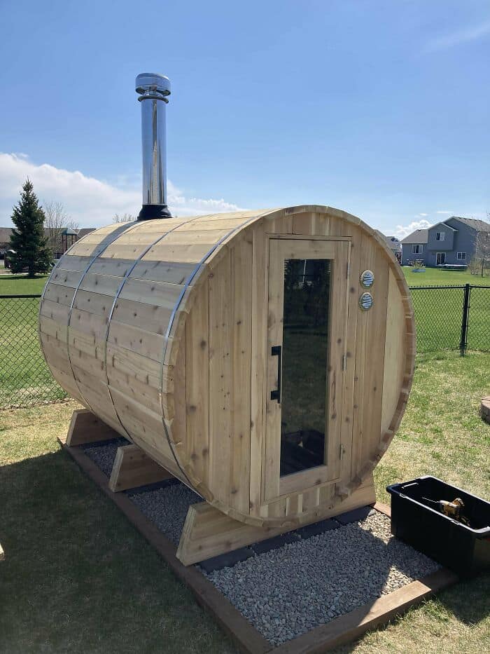Outdoor cedar barrel sauna with a glass door and metal chimney pipe. A great example of amazing woodworking.