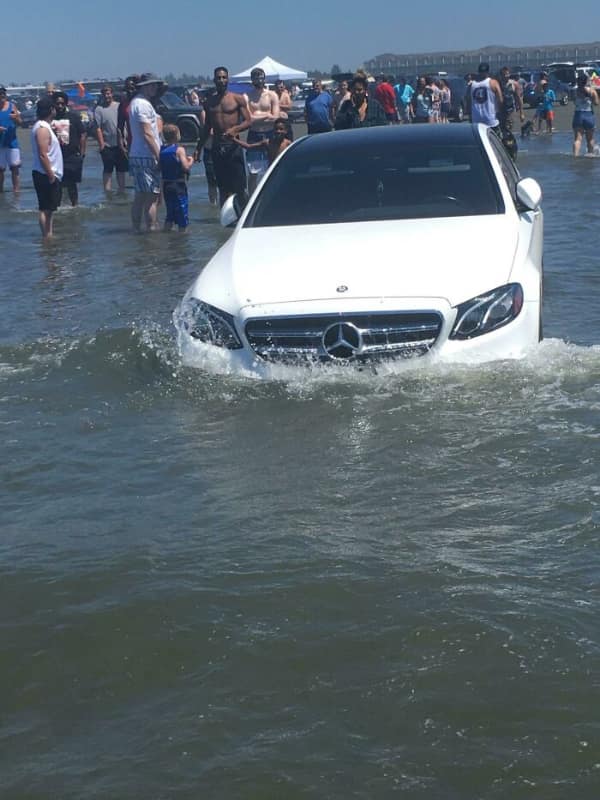 driving fail: A white Mercedes-Benz sedan partially submerged in ocean water on a crowded beach as waves wash against the hood
