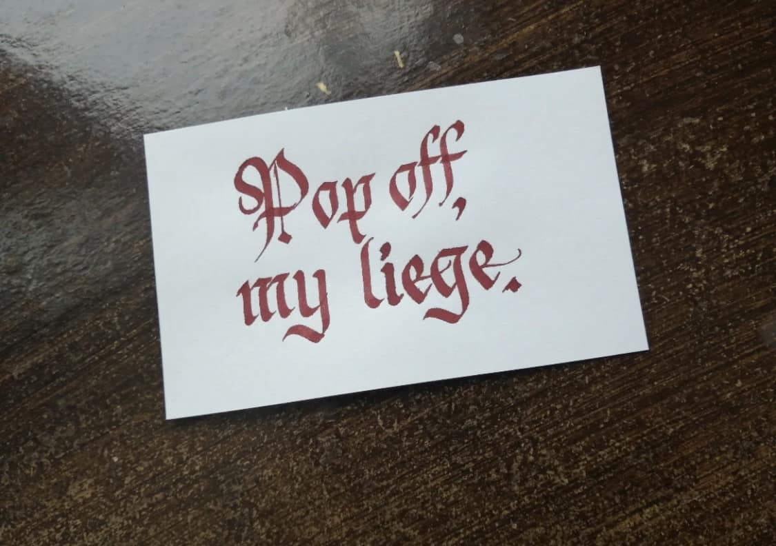 A fantastic artistic vintage meme showing a piece of parchment paper with beautiful, elegant gothic red calligraphy spelling out the hilariously modern internet slang phrase, "Pop off, my liege.".