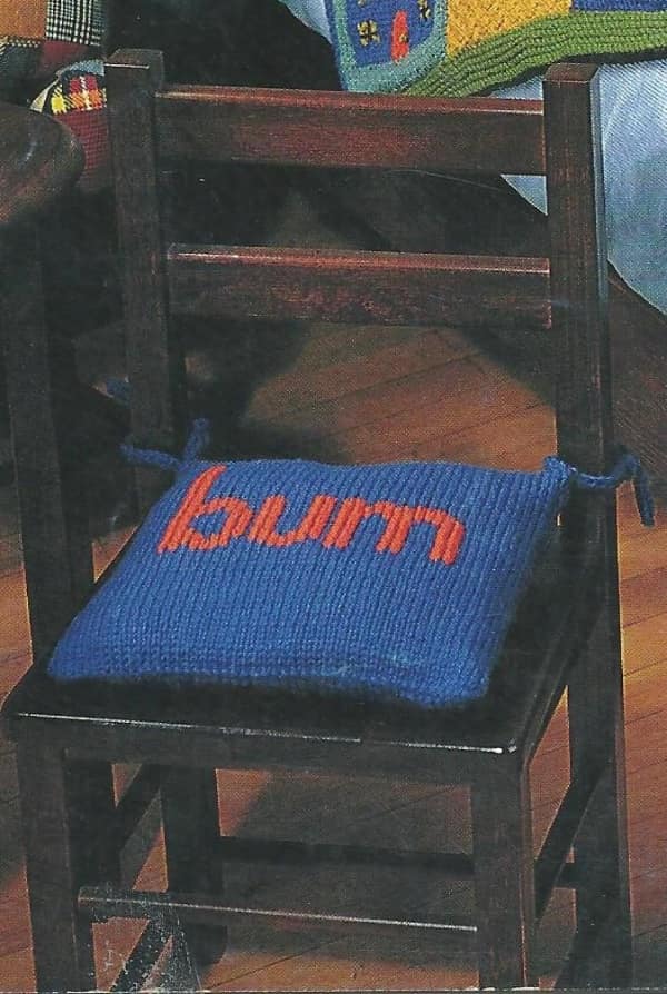 A blue creative knitting project chair cushion with the word "bum" knitted into it in red letters.