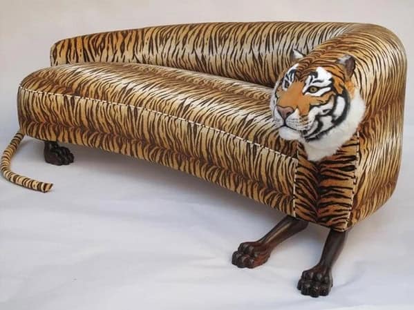 A weird furniture chaise lounge upholstered in tiger print fabric featuring a realistic 3D tiger head and tail attached to the ends.