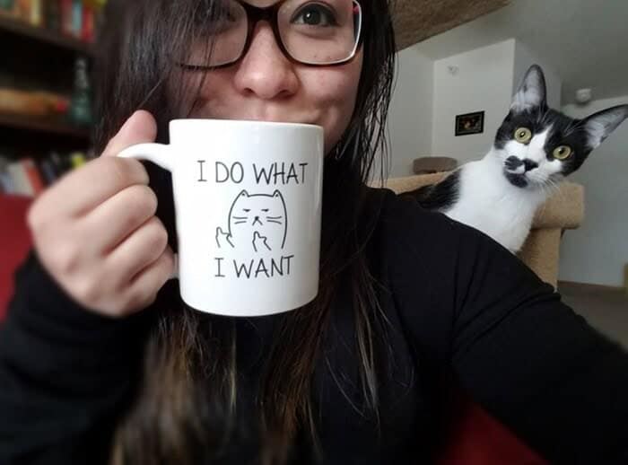 A hilarious funny animal photobomb where a black and white cat stares intensely from the background while its owner holds a mug reading "I do what I want."