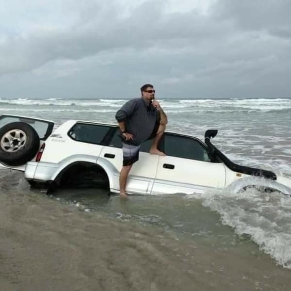 A confident but disastrous bad day photo showing a man posing proudly with his foot resting on his white SUV, which is hopelessly sunk deep into the wet sand and ocean waves on a beach.